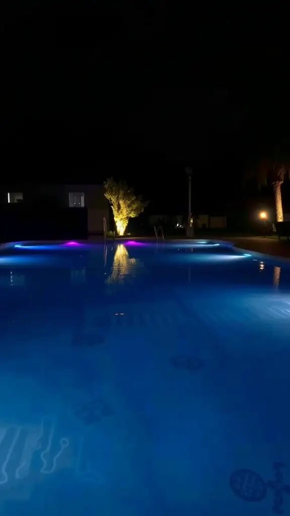 A nighttime view of a backyard pool illuminated with colorful underwater lights, surrounded by softly lit trees.